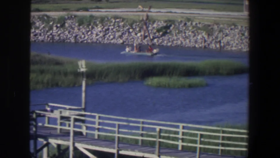 1971 Saint Paul home-movie frame showing a boat near the far stone bank, with reeds and white pier railing framing the water.