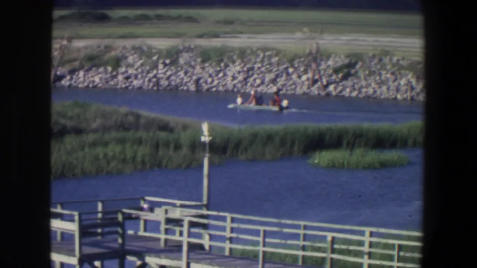 1971 Saint Paul frame showing a small motorboat with distant people passing reed beds below a stone embankment and grassy shore.
