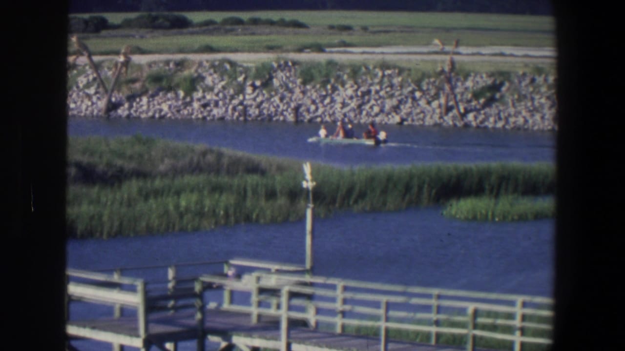 Small motorboat crossing blue water beyond a wooden dock and marsh grass.