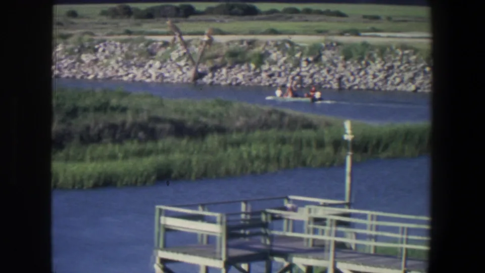 Home-movie frame of a motorboat crossing a lake in Saint Paul, Minnesota, with dock railings and marsh grass below.
