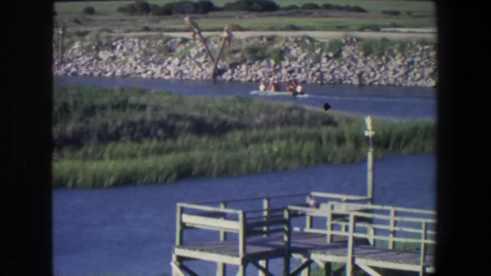 Small motorboat with passengers on blue lake water, framed by a wooden dock, reeds, and a rocky Saint Paul shoreline.