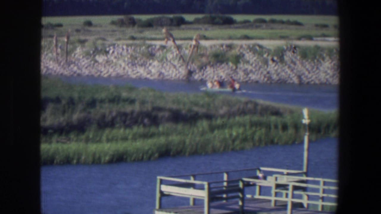 Small motorboat on blue water near marsh grass, a rocky shoreline, and a wooden dock railing.