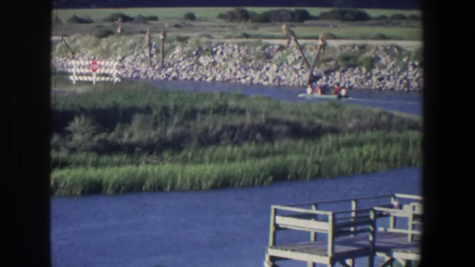 1971 Saint Paul waterway view with marsh grass, rocky bank, scattered fence posts, a small red sign, and dock railing at the lower edge.
