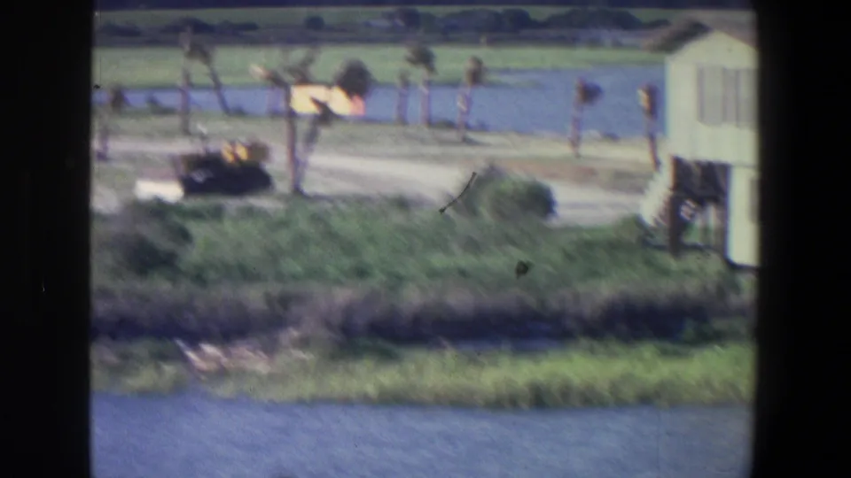 1971 Saint Paul home-movie frame showing a yellow bulldozer by a pale waterside structure, posts, grass, and water in the foreground.