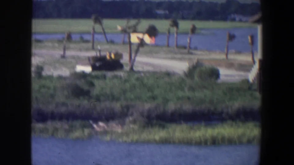 1971 Saint Paul waterfront frame with a bulldozer, shoreline work area, posts, grass, open water, and part of a pale structure at right.