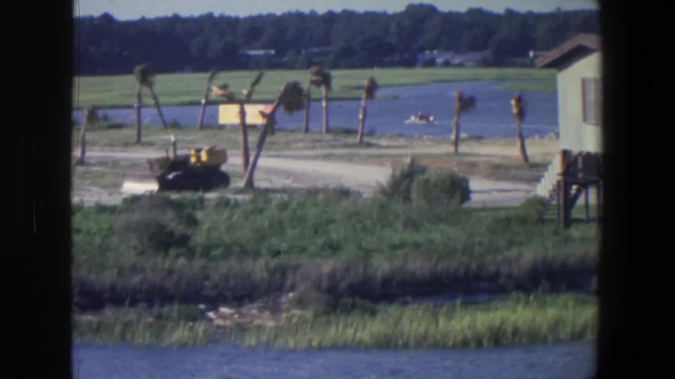 1971 Saint Paul waterfront view with dark frame edges, a bulldozer near palms and marsh grass, and a small boat moving across the water.