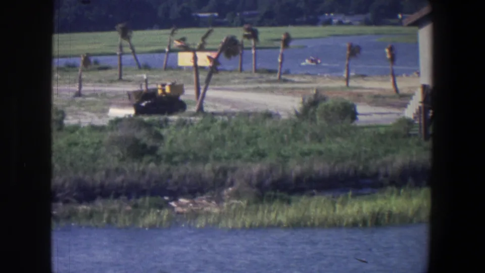 1971 Saint Paul home-movie frame with a soft, grainy view of a bulldozer, palms, marsh grass, water, and a distant boat.