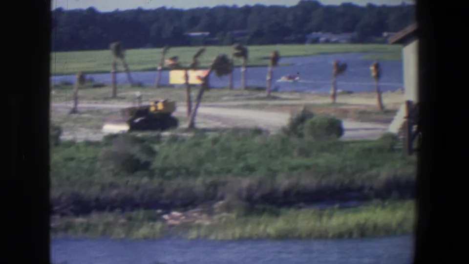 1971 Saint Paul waterfront frame showing a bulldozer near the shore, palm trees, marsh grass, dark frame edge, and tiny distant boats.