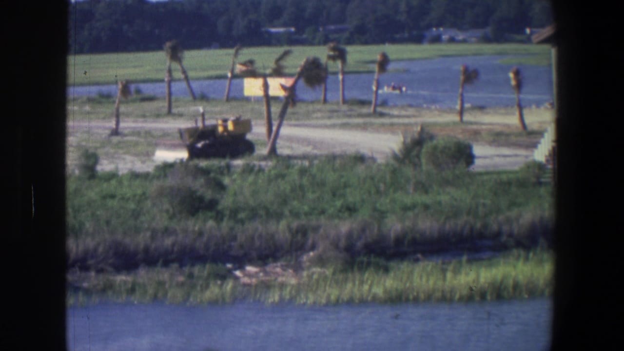 1971 Saint Paul 8mm frame showing blue lake water, grassy shore, a dirt path, small trees, and a yellow bulldozer beyond the reeds.