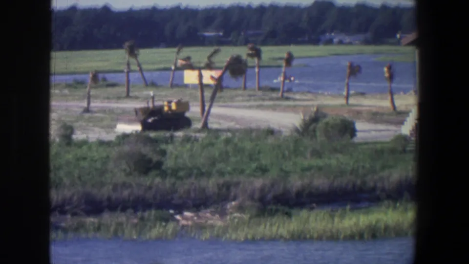 1971 Saint Paul lakeside view with reeds in the foreground, a dirt path, a yellow bulldozer, and small trees against open grass and water.
