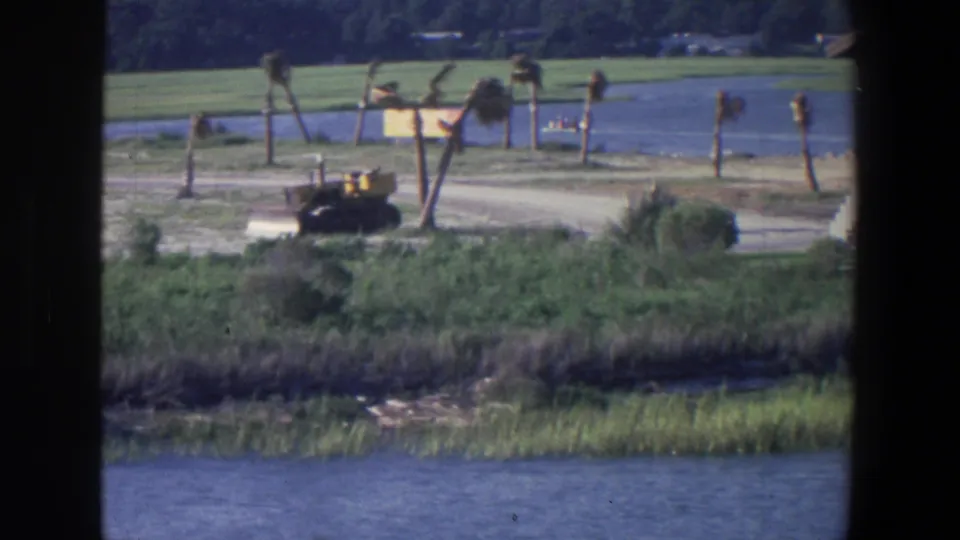 1971 Saint Paul home-movie frame showing a waterfront strip with reeds, green grass, a dirt path, small trees, and a yellow bulldozer.