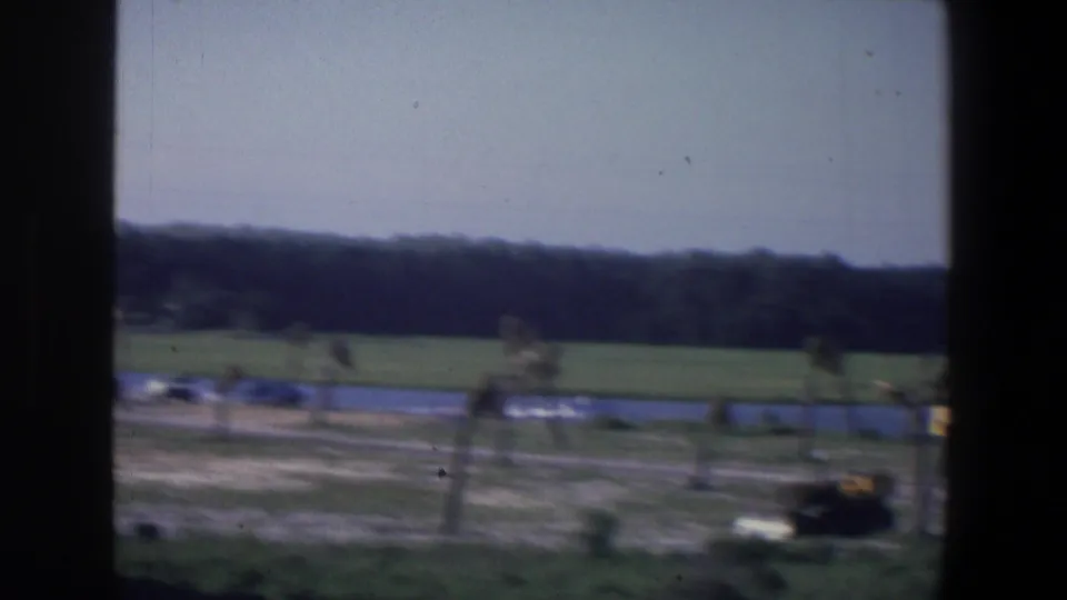 Darker 1971 Saint Paul 8mm frame with a blurred lakeside view, open grass, distant tree line, and a yellow bulldozer near the right edge.