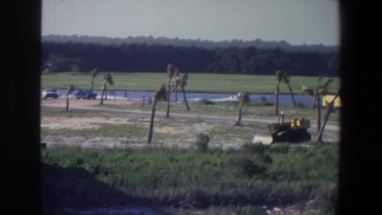 Distant yellow tractor near a dirt road, with water, grass, open field and tree line beyond.