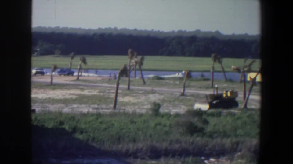 1971 8mm view near Saint Paul showing a yellow tractor by a dirt road beside water, with fields and trees in the distance.