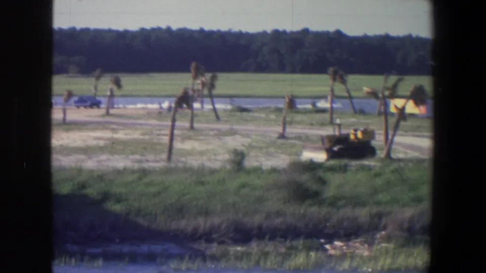 1971 Saint Paul-area rural home-movie frame with a yellow tractor, dirt road, water and broad green field under daylight.