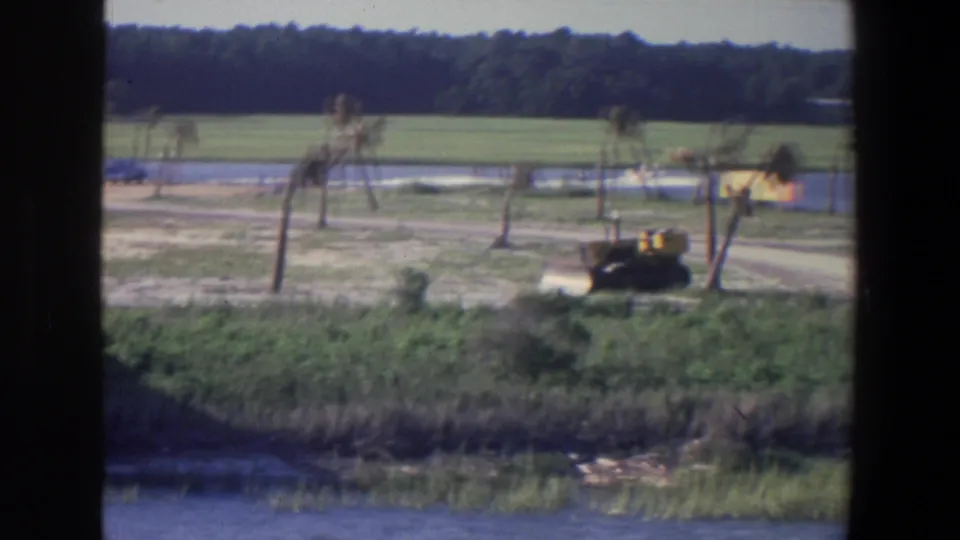 Soft 1971 8mm frame from Saint Paul showing a tractor near water and a dirt road, with open fields and trees beyond.