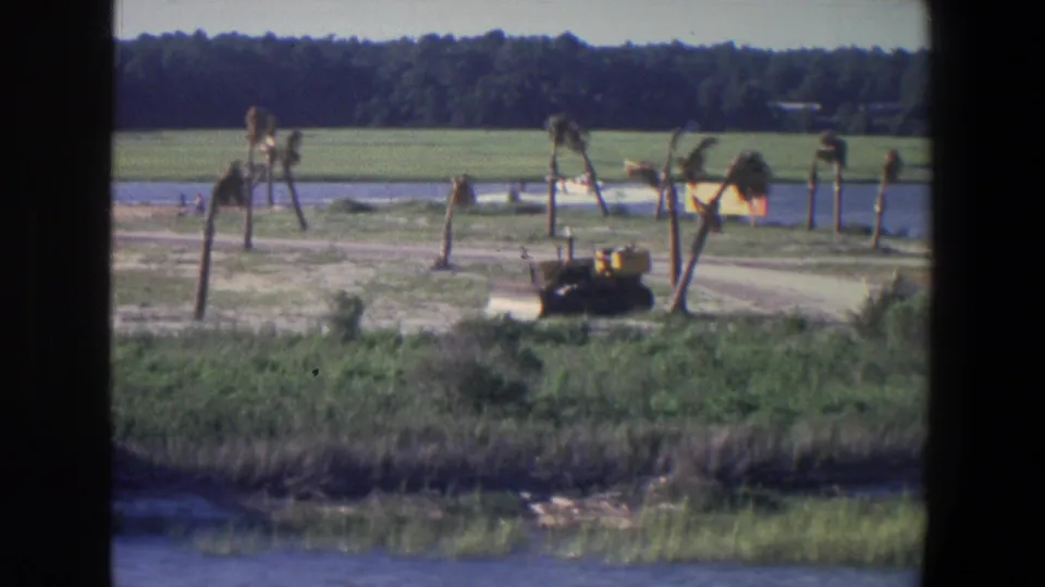 1971 Saint Paul rural frame showing a yellow tractor by a road near water, with green field and wooded horizon behind it.