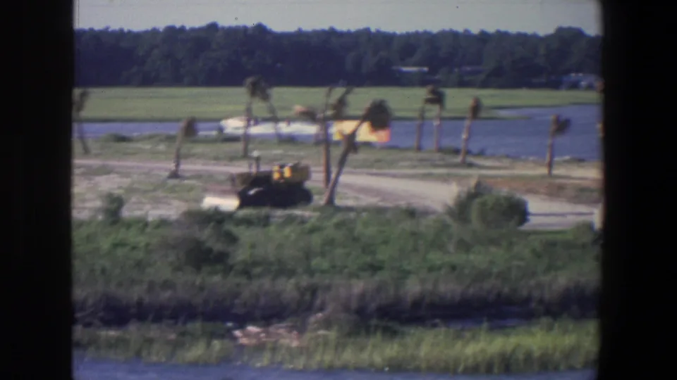 1971 Saint Paul 8mm frame showing a distant white speedboat near shore, with open grass, palm-like trees, and a yellow construction vehicle.