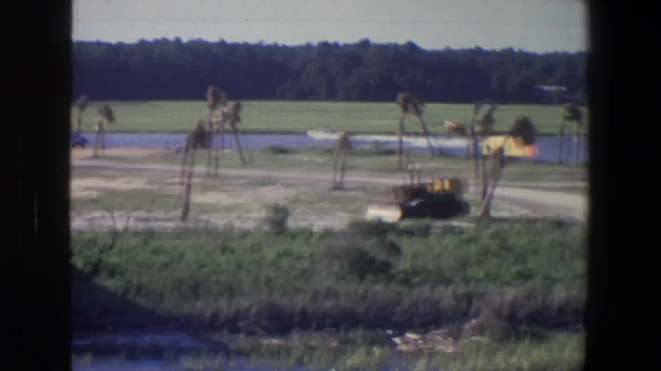 1971 Saint Paul lakeside frame with a distant white boat on the water, sparse trees along shore, open grass, and a yellow construction vehicle.