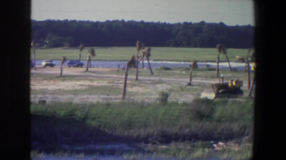 1971 Saint Paul 8mm frame showing cars along the lakeside, a distant boat area, sparse palm-like trees, and a yellow construction vehicle.