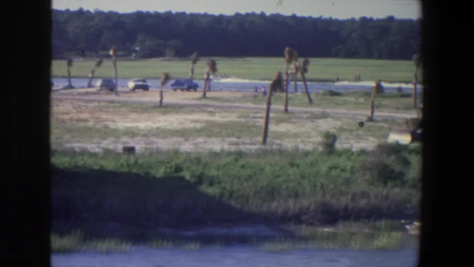 1971 Saint Paul home-movie frame of a distant white boat and cars beside the lake, with open grass, scattered shoreline trees, and heavy gate borders.