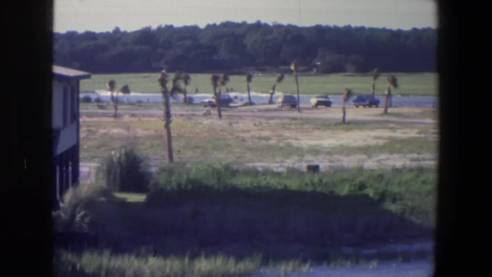 1971 8mm frame identified as Saint Paul, Minnesota, showing a waterside structure, grassy bank, parked cars, and palm-like trees beyond the river edge.
