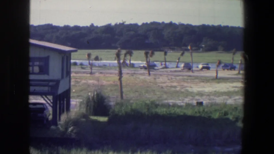 Frame from the 1971 riverside home movie shows a small elevated structure, cars along a road, open grass, and a wooded background.