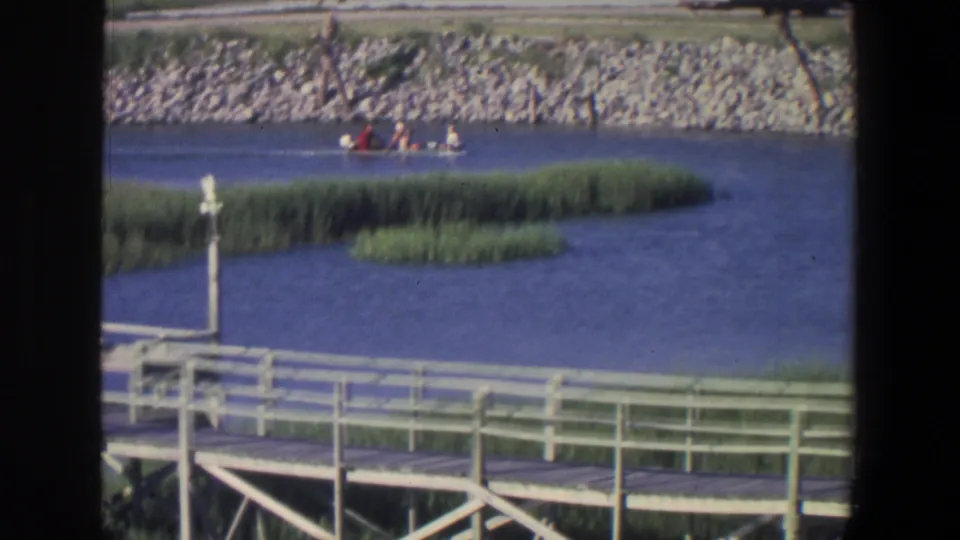 1971 riverside frame shows a small boat carrying people near reeds, with a dock or walkway railing in the foreground and a rocky bank behind.