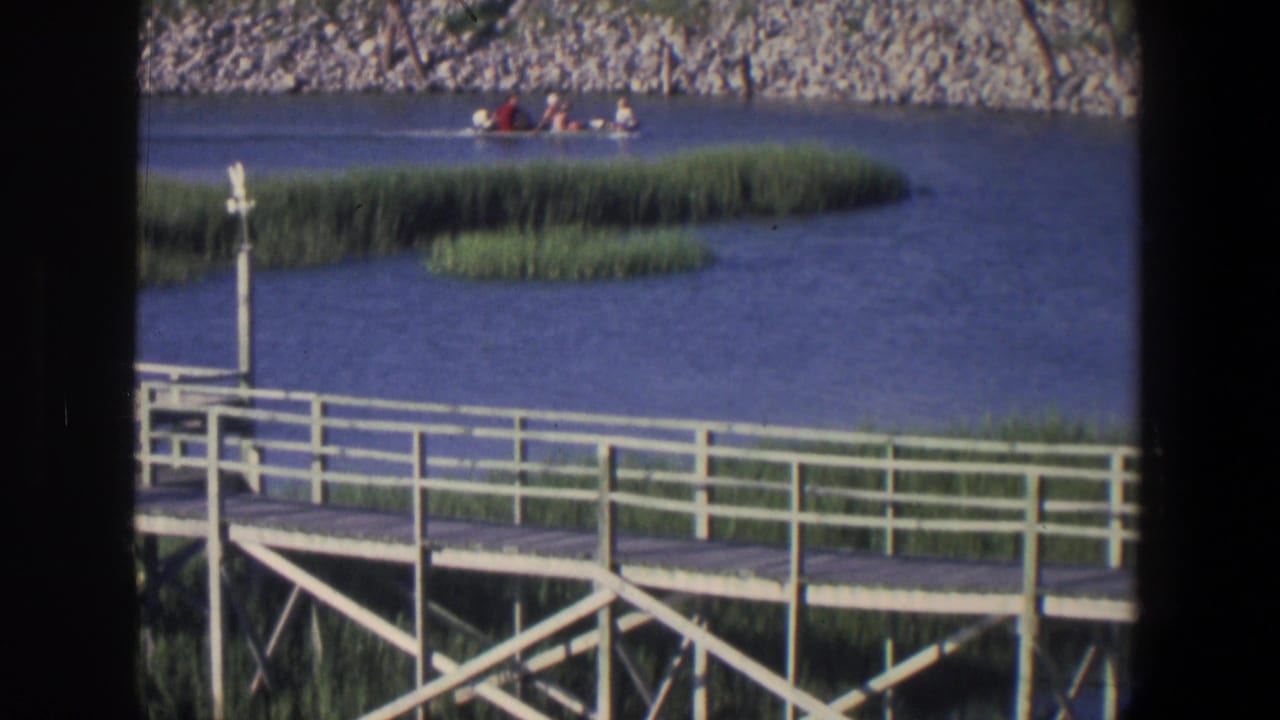 1971 Saint Paul home-movie frame showing a small motorboat with passengers beyond a white dock, marsh grass, and rocky shore.