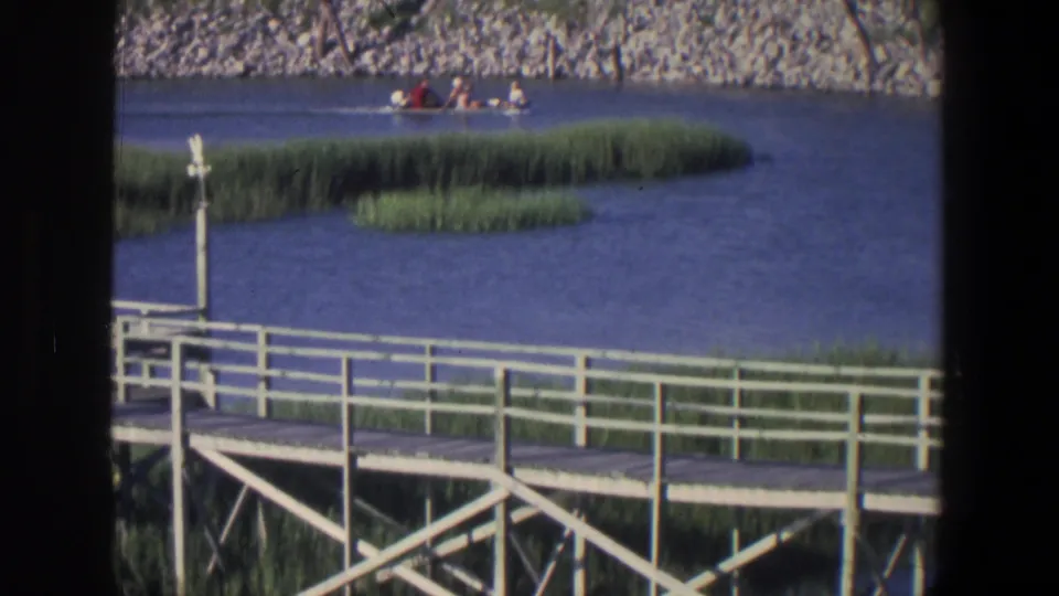 1971 Saint Paul home-movie frame showing a small motorboat with passengers beyond a white dock, marsh grass, and rocky shore.