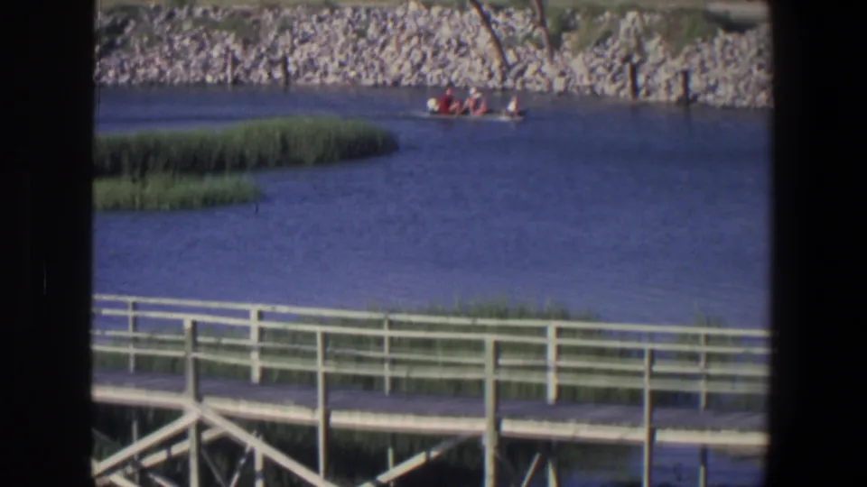1971 Saint Paul waterfront frame with a small passenger motorboat near the rocky shoreline, seen over dock rails and reeds.