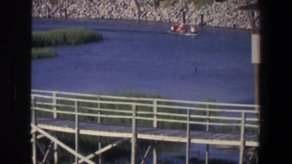 1971 Saint Paul home-movie frame showing a small boat on calm water behind a wooden boardwalk and riverside reeds.
