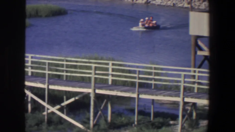 1971 home-movie still of several people in a small boat moving across a calm river near a white-railed boardwalk.