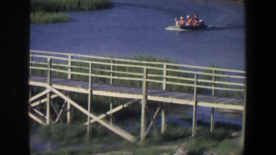 Small boat with passengers on a Saint Paul river, framed by marsh vegetation and a wooden boardwalk in 1971 8mm film.