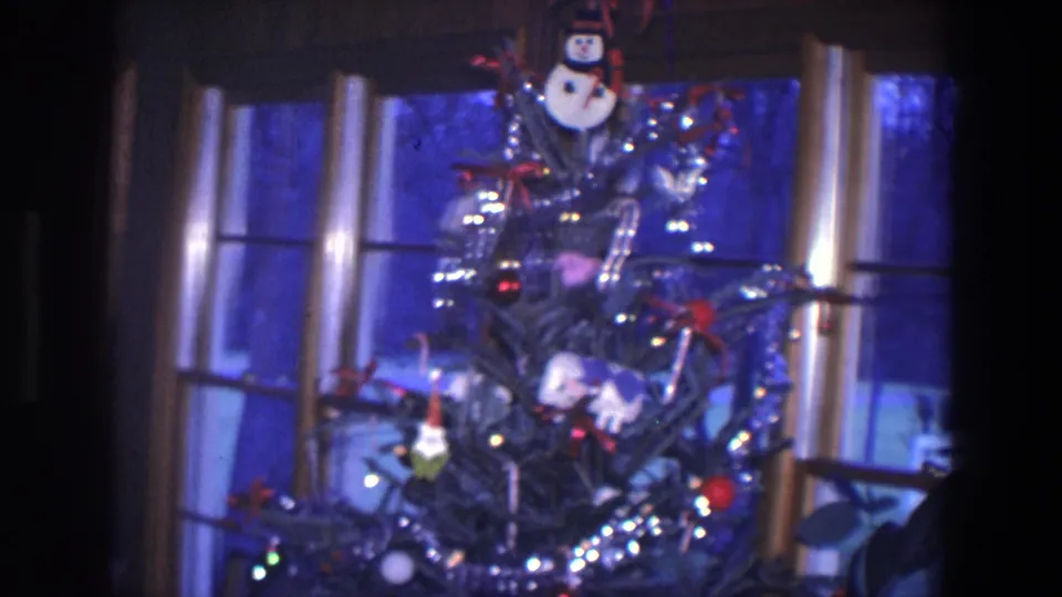 1971 Saint Paul home-movie frame showing a Christmas tree with tinsel, lights, ornaments, and a snowman decoration by a dark window.