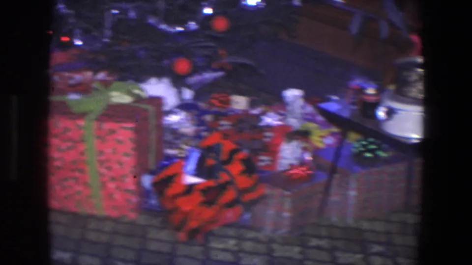 1971 home-movie view of Christmas gifts beneath a tree, with tinsel, ornaments, patterned carpet, and a table entering the frame.