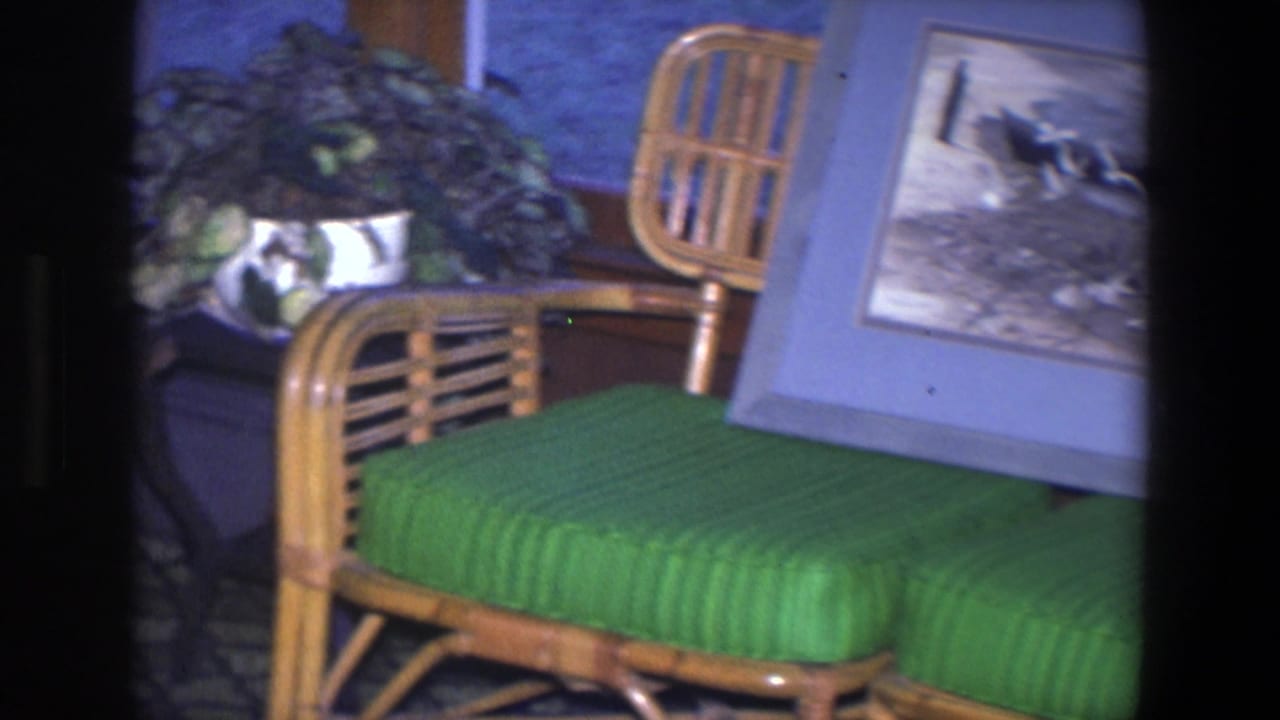 1971 Saint Paul home-movie frame showing framed animal artwork on wicker seating with a green cushion in a private interior.