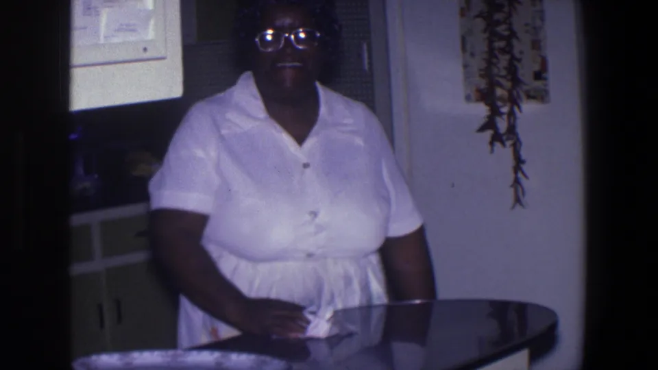 African American woman in glasses and a white dress stands by a kitchen table in a 1971 Saint Paul home-movie kitchen scene.