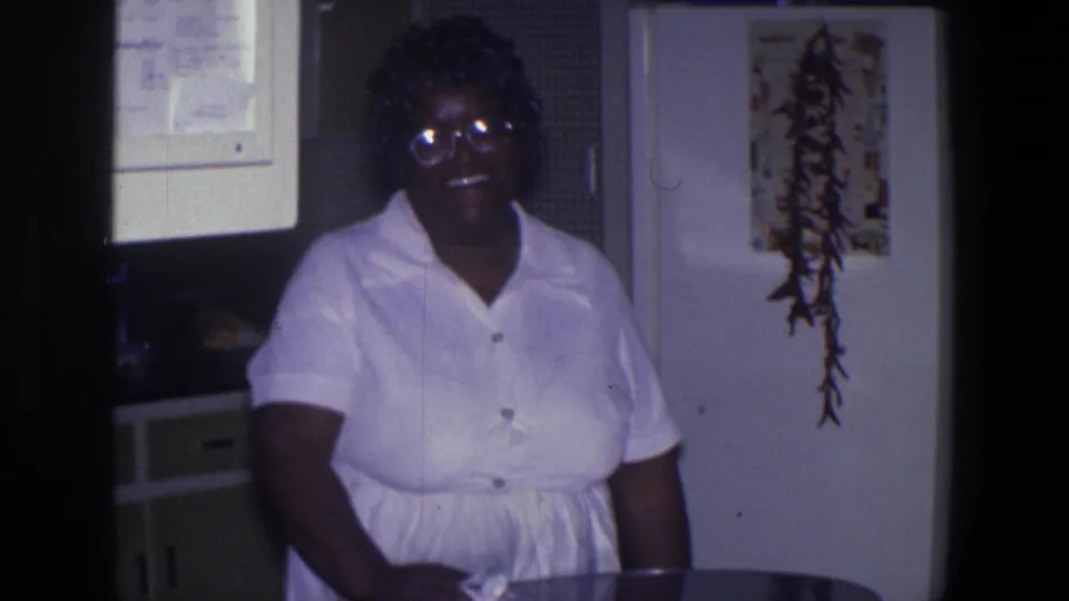 Smiling African American woman in glasses stands beside a kitchen table, with a refrigerator and hanging plant visible in the Saint Paul home interior.