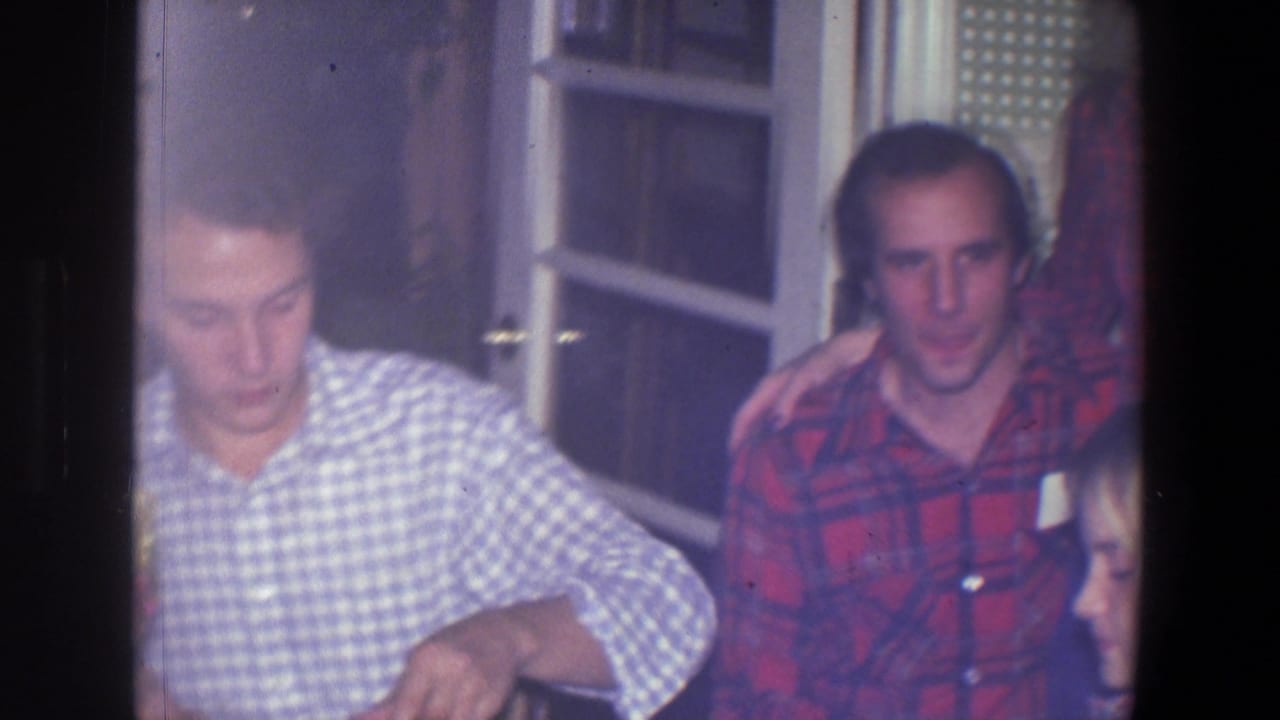 In a Saint Paul 1971 home-movie frame, a young man in a blue and white checkered shirt sits at a table near windowed doors.