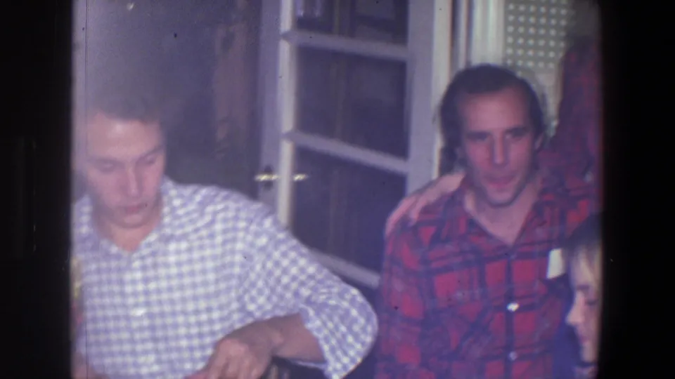 In a Saint Paul 1971 home-movie frame, a young man in a blue and white checkered shirt sits at a table near windowed doors.
