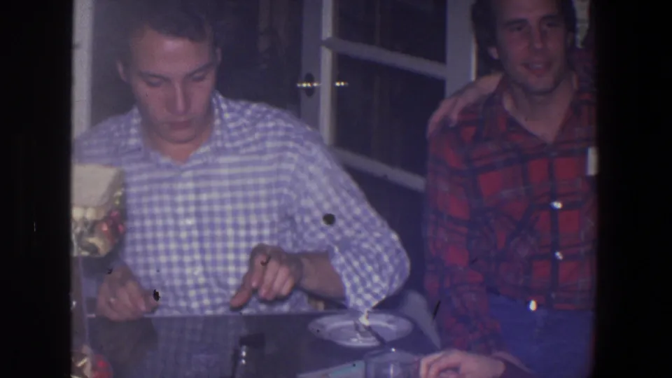 Two young men sit at a table inside a Saint Paul home, with a plate and cup visible in soft 1971 8mm footage.
