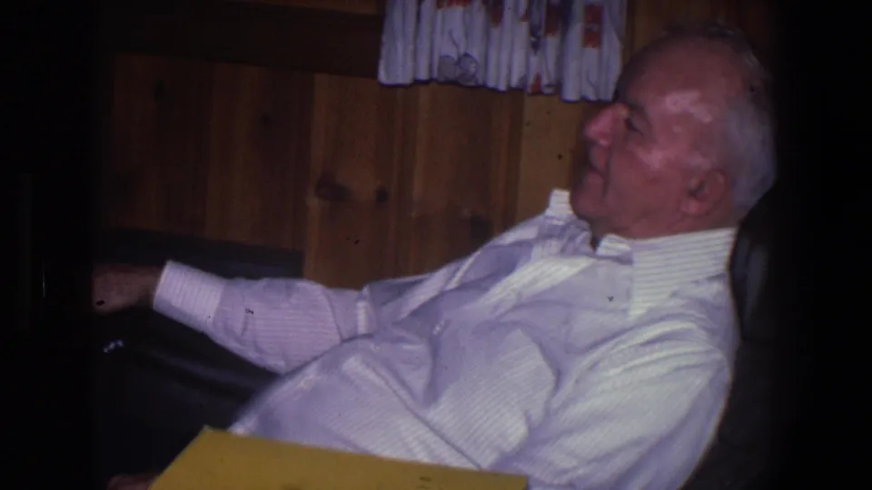 An older man remains seated in a chair, appearing to smile or speak, with wood paneling and curtains behind him in the home.