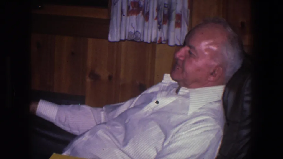 A 1971 home-movie view shows the elderly man reclining in the chair beneath a patterned curtain and wood wall.