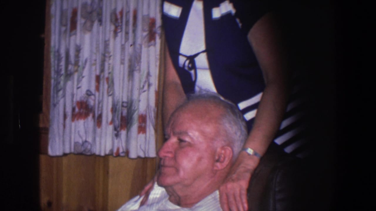 In a 1971 Saint Paul home interior, an older man sits while a woman stands behind him with her hands near his shoulders.