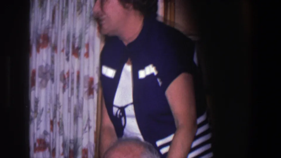 A woman in a dark vest with white trim smiles while standing behind an older seated man in a Saint Paul home-movie scene.