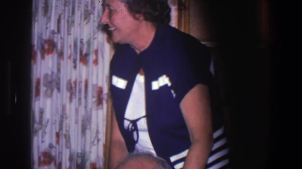 The standing woman remains near the floral curtains, with part of the older seated man visible at the lower edge of the frame.