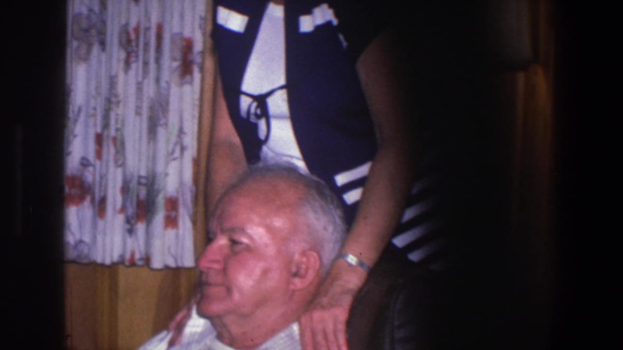 Elderly man seated while a woman stands behind him with hands near his shoulders.