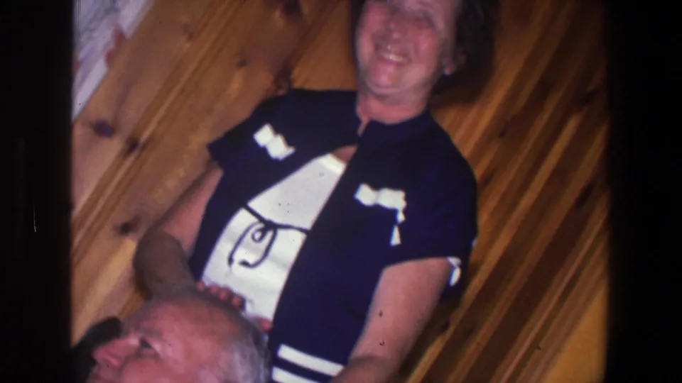 A smiling woman in a dark blue and white dress stands behind an elderly man in a wood-paneled Saint Paul home interior.
