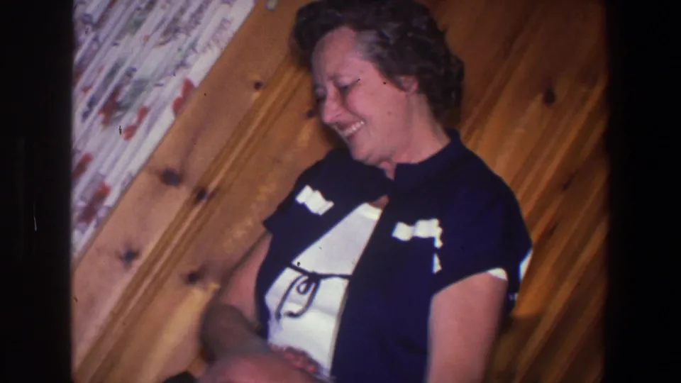 The woman laughs as she leans near the seated elderly man, with wood paneling and curtains visible in the 1971 home-movie frame.